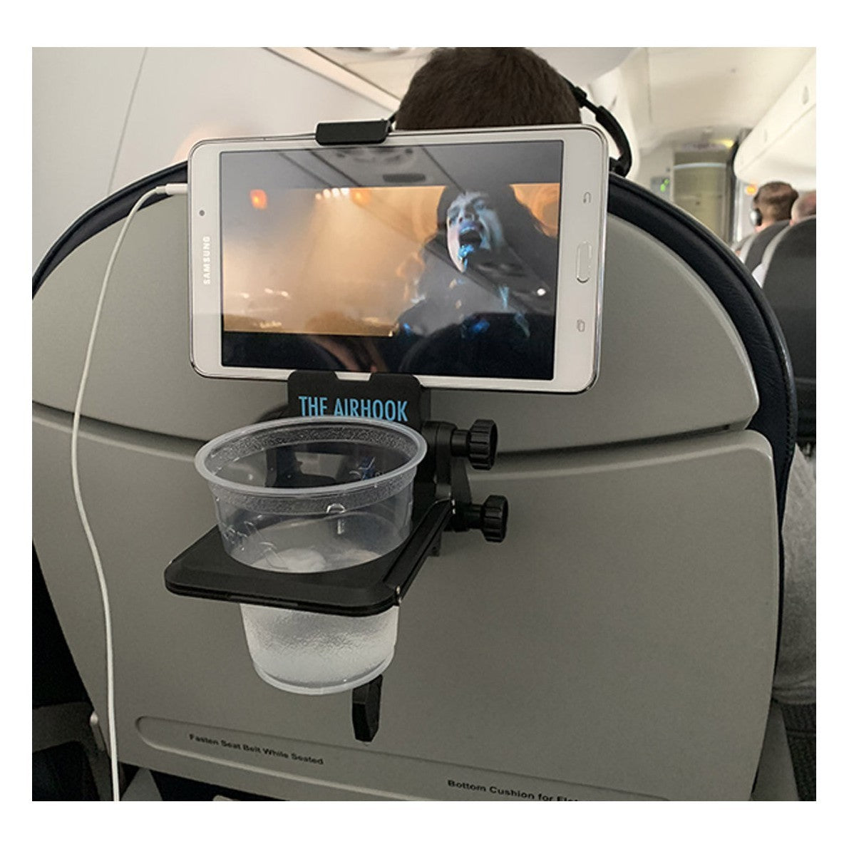 The AIRHOOK shown with a smartphone playing a movie with a plastic cup and beverage while attached to the back of an airplane seat.